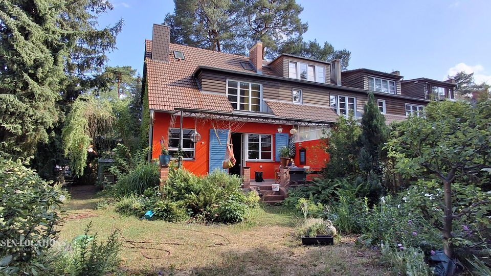 Rückansicht eines mehrstöckigen Reihen- bzw. Doppelhauses mit rotem Erdgeschoss, blauem Eingang und braunem Satteldach mit Gauben und großen Fenstern. Davor liegt ein verwachsener Garten mit Rasenfläche, Sträuchern, Bäumen und einer kleinen Terrasse mit Treppenstufen.