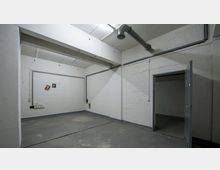 Kleiner, leerer Raum mit weiß gestrichenen Ziegelwänden, einem kahlen Betonboden und sichtbaren Rohrleitungen an der Decke. Eine geöffnete Metalltür führt in einen angrenzenden Raum, und an einer Wand hängen zwei kleine Bilder.