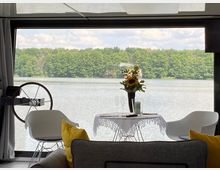 Blick von innen durch eine große Fensterfront auf einen See mit bewaldetem Ufer im Hintergrund. Im Vordergrund stehen ein gedeckter runder Tisch mit Sonnenblumen und zwei weißen Stühlen sowie ein Steuerrad auf der linken Seite.