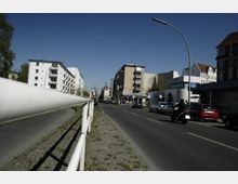 Straßenszene mit Bürgersteig und Metallgeländer im Vordergrund, Mehrfamilienhäuser auf der linken Seite und Geschäfte sowie ein "U-Bahn"-Schild auf der rechten Straßenseite; parkende Autos und eine Motorradfahrerin sind ebenfalls sichtbar. Der Himmel ist klar, ohne Wolken.