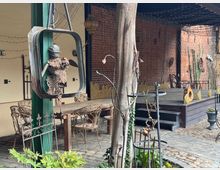 Hof im rustikalen Stil mit Backsteinwänden, Kopfsteinpflaster und Vintage-Möbeln. Im Vordergrund eine Metallskulptur, im Hintergrund eine kleine hölzerne Bühne und dekorative Elemente aus Metall und Holz.