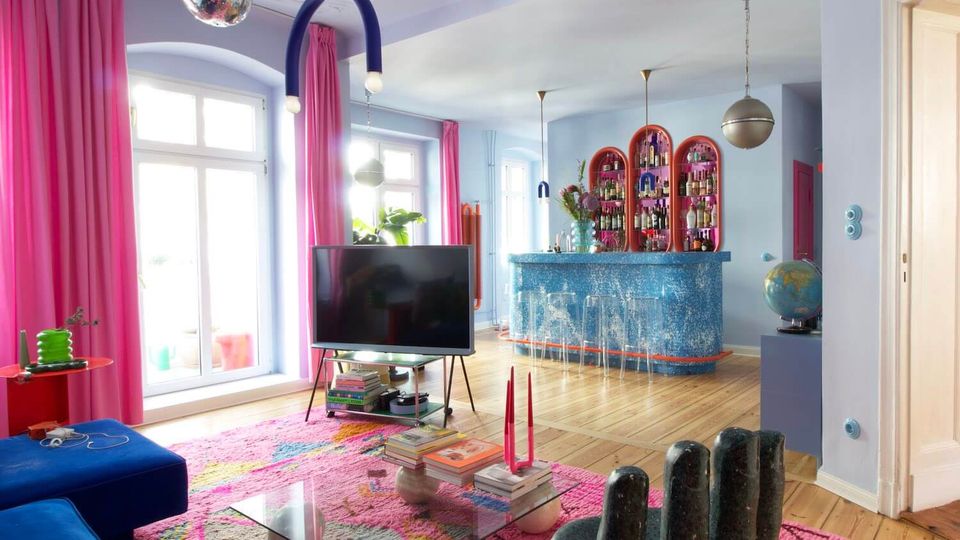 Bright, colorful living room with tall windows and pink curtains, light blue walls, and wood floors with a large patterned rug. A TV on a stand faces a blue sofa, with a glass coffee table in the center and an open bar area in the back featuring blue tiled counters and arched shelving.