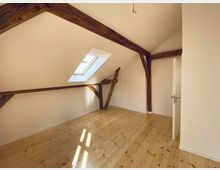 Dachgeschossraum mit weiß gestrichenen Wänden und sichtbaren Holzbalken an der Decke. Der Raum hat ein schräges Fenster, das natürliches Licht hereinlässt, sowie einen hellen Holzboden und eine geöffnete Tür auf der rechten Seite.