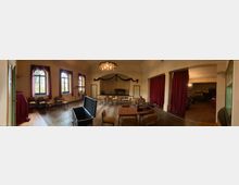 Ein historisch anmutender Veranstaltungsraum mit hohen Decken, großen Fenstern mit roten Vorhängen und einem Kronleuchter. Der Raum ist mit Stühlen, Tischen und einem Klavier ausgestattet, und rechts befindet sich eine Bühne mit roten Samtvorhängen.