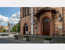 Außenansicht eines historischen Gebäudes aus rotem Backstein mit zwei großen Holztüren, einem Vordach und dekorativen Rundbögen. Vor der Kirche stehen Pflanzkästen, und im Hintergrund sind moderne Gebäude sowie Bäume zu sehen.
