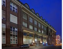 Großes Backsteingebäude mit großen weißen Fenstern und gewölbten Dachgauben, beleuchtet in der Dämmerung. Im Erdgeschoss sind Eingänge mit Vordächern, daneben geparkte Autos; im Hintergrund eine kaum belebte Straße mit weiteren Gebäuden.