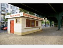 Ein kleiner Imbissstand unter einer erhöhten Eisenbahnbrücke in Berlin, mit weiß gefliester Fassade, rot-oranger Tür und einem Schild mit der Aufschrift „Bockwurst“ und „Berliner Currywurst“. Im Hintergrund sind Straßenszenen mit parkenden Autos, Bäumen und Gebäuden zu sehen.