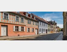 Eine gepflasterte Straße mit bunten historischen Fachwerkhäusern, deren Fassaden in Pastelltönen gestrichen sind. Im Hintergrund sind weitere Gebäude und ein Kirchturm unter einem klaren blauen Himmel zu sehen.
