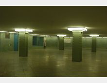 Großzügiger U-Bahn-Zugang mit grünen Metallstützen und rechteckigen Deckenleuchten, gefliesten Wänden in grün-blauen Tönen, sowie einer blauen Tür und Hinweisschildern an der Wand. Der Boden ist mit beigen Fliesen bedeckt.