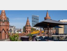 Blick auf die Oberbaumbrücke in Berlin mit ihren roten Backsteintürmen, im Hintergrund ein modernes Hochhaus und ein gelber Zug der Berliner U-Bahn. Im Vordergrund befindet sich eine Terrasse mit Pflanzen, Sitzmöbeln und einem Sonnenschirm.