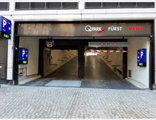Einfahrt zu einem Parkhaus mit der Beschilderung „Q-Park Fürst“ und „REWE“, flankiert von blauen Parkhinweisschildern mit der Anzeige „Frei“. Ein steiler, beleuchteter Auffahrtsweg führt ins Innere, mit Höhenbegrenzung und Verbotsschildern über der Einfahrt.
