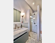 Modernes Badezimmer mit grauen Wänden und gläserner Duschkabine. Ein Waschtisch mit Marmor-Oberfläche und goldenem Wasserhahn steht davor, ergänzt durch geometrischen Fliesenboden und eine wandmontierte Lampe.