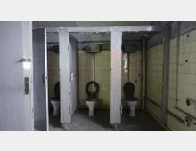 Abgenutzte öffentliche Toilettenanlage mit drei Kabinen, deren Türen teilweise fehlen oder offenstehen. Die Wände sind mit hellen Fliesen bedeckt, die teils verschmutzt und beschädigt sind; sichtbare Rohre und eine verblasste Graffiti-Markierung sind ebenfalls erkennbar.