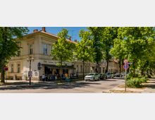 Straßenecke mit zweigeschossigem Altbau in heller Fassade und rotem Ziegeldach, im Erdgeschoss ein kleiner Laden mit Vordach. Entlang der von großen, belaubten Bäumen gesäumten Straße stehen parkende Autos und Verkehrsschilder am Gehweg.