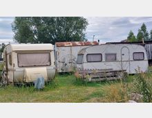Zwei ältere Wohnwagen stehen auf einer Wiese mit hohem Gras, neben ihnen ein Fahrrad und Holzbretter. Im Hintergrund sind ein verrosteter Container, Bäume und Teile von Gebäuden zu sehen.