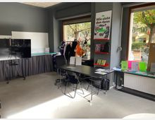 Ein moderner Workshopraum mit grauen Wänden, großen Fenstern mit Blick auf eine Baumreihe und mehreren Tischen und Stühlen. An der Wand hängt ein Plakat, daneben stehen ein Fernseher auf einem Rollständer, ein Whiteboard und Regale mit Büchern und Materialien.