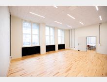 Heller, leerer Innenraum mit hellem Holzfußboden, hohen Fenstern entlang einer Wand und schwarzen Heizkörpern darunter. Abgehängte Decke mit langen LED-Lichtleisten sowie eine Türöffnung zu einem angrenzenden Raum mit Tischen und Stühlen.