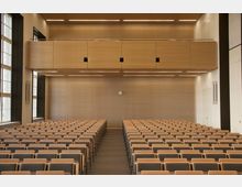 Ein moderner Hörsaal mit mehreren Reihen gepolsterter Sitze und einem schlichten Holzdesign an den Wänden und der Decke. Große Fenster auf der linken Seite lassen Tageslicht herein, und eine Uhr ist zentriert an der Rückwand angebracht.