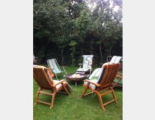 Ein Garten mit mehreren Holz- und Liegestühlen, die um eine runde Feuerstelle herum angeordnet sind; im Hintergrund stehen dichte grüne Bäume und Sträucher.