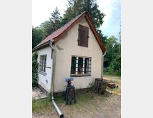 Kleines Gebäude mit hell verputzten Wänden, rotem Satteldach und einer Holztür im oberen Giebel. Vor dem Haus stehen eine Werkbank mit Schraubstock und ein Baumstumpf, im Hintergrund befinden sich Bäume und Wiese.