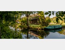 Holzhaus mit schrägem Dach an einem kleinen, baumbestandenen Fluss; im Vordergrund ein mit grünem Planenstoff bedecktes Boot am Ufer, umgeben von dichtem Laub und gepflegtem Rasen.