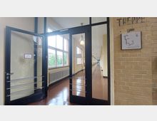 Ein heller Flur in einem Gebäude, mit gelb gestrichenen Wänden und großen Fenstern auf der linken Seite, die Tageslicht hereinlassen. Im Vordergrund befindet sich eine Glastür mit schwarzem Rahmen, rechts eine Backsteinmauer mit einem Treppenschild und einem Grundriss des ersten Obergeschosses.