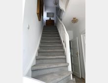 Eine Innentreppe mit grauem Teppichbelag, weißem Geländer und leicht geschwungener Form führt nach oben zu einem kleinen Bereich mit einer Holzkommode und einem Spiegel an der Wand. Zwei dekorative Kronleuchter hängen von der Decke, und Türen sind links und rechts im unteren Bereich sichtbar.