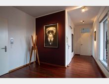 Flur eines modernen Wohnbereichs mit Holzboden, weiß gestrichenen Wänden und einem dunkelvioletten Akzentbereich. An der Akzentwand hängt ein gerahmtes Bild eines Affen mit Sonnenbrille und Anzug, daneben steht ein minimalistischer Holz-Kleiderständer.