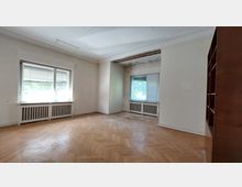 Ein leerer Raum mit abgenutztem Parkettboden, großen Fenstern mit Jalousien und Heizkörpern darunter. Die Wände sind weiß, teilweise beschädigt, und an der Decke ist ein Abschnitt mit freiliegendem Putz sichtbar; auf der rechten Seite befindet sich ein eingebautes Holzregal.