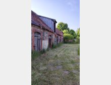 Eine ländliche Szene mit alten Ziegel- und Feldsteingebäuden, teilweise mit Holzverkleidung. Davor befindet sich eine gemähte Wiese, umgeben von Bäumen und Sträuchern im Hintergrund.