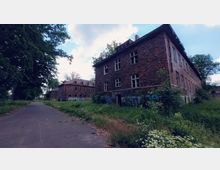 Ziegelgebäude in einem verwilderten Außenbereich, mit teilweise zerstörten Fenstern und Graffiti an den Wänden; eine asphaltierte Straße führt an den Gebäuden vorbei, umgeben von Bäumen und dichter Vegetation.