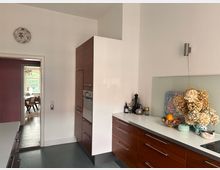 Moderne Küche mit Holzschränken und weißer Arbeitsplatte, dekoriert mit getrockneten Blumen und Obstschale. Im Hintergrund führt eine Tür zu einem Esszimmer mit Holzboden und großen Fenstern.