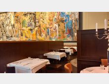 Elegantes Restaurant mit gedeckten Tischen, weißen Tischdecken und dunklen Holzmöbeln. Eine große, farbenfrohe Wandmalerei mit abstrakten und figürlichen Motiven dominiert den Raum.