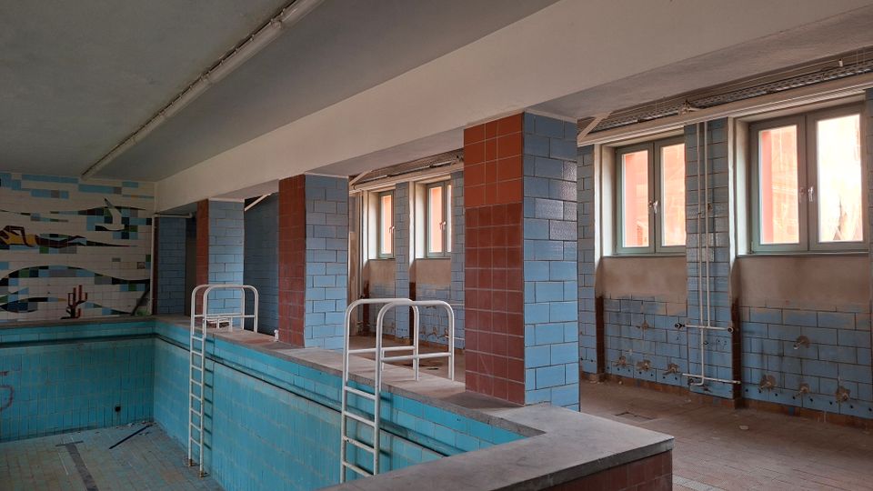 Verlassenes Hallenbad mit leerem, blau gefliestem Becken und mehreren Metallleitern entlang des Beckenrands. Blaue und rotbraune Fliesenpfeiler tragen die Decke; an der rechten Wand befinden sich Fenster, freiliegende Rohrleitungen und abblätternde Fliesen.
