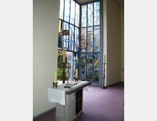 Moderne Kapelle mit einem schlichten Steinaltar, der mit einem weißen Altartuch und Kerzen dekoriert ist. Im Hintergrund sind hohe, farbige Glasfenster mit abstrakten blau- und gelbtönigen Mustern zu sehen.