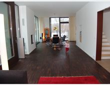 Ein moderner Wohnraum mit dunkelbraunem Holzboden, einer großen Fensterfront im Hintergrund mit Blick ins Freie, und einer Essecke mit schwarzen Stühlen. Rechts befindet sich eine weiße Treppe, und links sind eine Tür sowie große Glasflächen sichtbar. Ein Kind läuft durch den Raum.