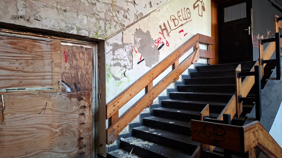 Verlassenes Treppenhaus in einem Gebäude mit dunklen Stufen und Holzgeländern, die zu einer geschlossenen Tür im oberen Stockwerk führen. Abblätternder Putz, Graffiti an der Wand und eine mit Sperrholz vernagelte Öffnung prägen den heruntergekommenen Innenraum.