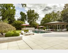 Moderne Gartenlandschaft mit einem großzügigen Pool, umgeben von gepflegten Rasenflächen und Büschen. Auf der Terrasse stehen Liegen und Sitzgruppen mit roten Sonnenschirmen, daneben ein gläsernes Pavillongebäude mit Tischen und Sitzgelegenheiten.