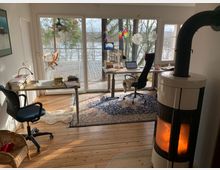 Ein heller Arbeitsraum mit Holzboden, ausgestattet mit zwei Schreibtischen, Bürostühlen, einem Teppich und einem Kaminofen mit sichtbarem Feuer. Große Fenster und eine Glastür bieten Ausblick auf eine Terrasse mit Holzmöbeln, umgeben von Bäumen und einem See im Hintergrund.