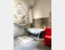 Badezimmer mit einer freistehenden Badewanne und Duschkopf, einer weißen Wand mit Spuren von Abnutzung, einem Waschbecken mit Spiegel in goldener Rahmenform und einem roten Polstersessel. Der Boden ist mit grauen Fliesen ausgelegt, und vor dem Waschbecken liegt eine weiße Badematte.