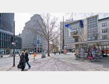 Städtische Straßenszene mit modernen Hochhäusern, darunter Gebäude mit Allianz-Logo, und einem kleinen Kiosk mit Souvenirs im rechten Vordergrund. Kahle Bäume und Menschen, die über eine breitere Pflasterstraße flanieren, prägen die Szenerie.