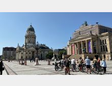 Weitläufiger Platz in Berlin mit Blick auf den Französischen Dom links, ein Barockgebäude mit Kuppel, und das Konzerthaus Berlin rechts, mit Säulenfassade und Statuen verziert. Im Vordergrund stehen Menschen mit Fahrrädern, der Platz ist von klassizistischer Architektur umgeben.