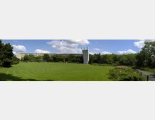 Weitläufige Grünfläche mit gepflegtem Rasen, umgeben von Bäumen und Sträuchern. Im Hintergrund ein modernes Gebäude und ein auffälliges, gebogenes Denkmal aus Beton, das aus der Landschaft herausragt.