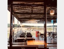 Überdachte Terrasse eines Restaurants oder Cafés mit Lichterketten an der Decke, mehreren Tischen mit Stühlen und Blick auf eine offene Landschaft mit Gras und Bäumen im Hintergrund.