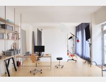 Heller Raum eines Fotostudios mit Holzfußboden, einem Schreibtisch mit Monitor und Stuhl, Regalen mit Aufbewahrungsboxen und Fotoausrüstung wie Lichtstativen, einem großen weißen Hintergrund und Vorhängen vor den Fenstern.