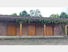 Backsteingebäude mit drei hölzernen Doppeltüren und Rankpflanzen, die entlang des Dachvorsprungs wachsen. Vor dem Gebäude befindet sich ein gepflasterter Bereich mit vereinzeltem Unkrautbewuchs, im Hintergrund sind Bäume zu sehen.