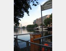 Uferbereich in Berlin mit Blick auf einen Fluss, umgeben von Bäumen und modernen Gebäuden. Im Vordergrund ein Geländer mit einer orangen Metalldose und einem Schild mit der Aufschrift „Historischer Hafen Berlin“, im Hintergrund liegen Boote am Ufer.