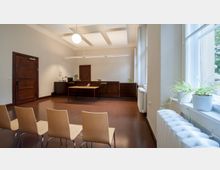 Ein minimalistischer Verwaltungsraum mit einer schlichten Holztheke im Hintergrund, einem kleinen Tisch in der Mitte und einer Reihe von Stühlen im Vordergrund. Große Fenster an der rechten Seite lassen viel Tageslicht herein und sind mit Blumentöpfen dekoriert.