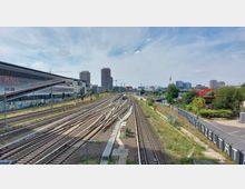 Blick auf ein Bahngleisfeld mit mehreren Gleisen, flankiert von modernen Gebäuden, darunter eine große Halle auf der linken Seite. Im Hintergrund sind Baukräne, Hochhäuser und der Berliner Fernsehturm zu sehen.