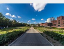 Breiter, gepflasterter Weg in einem parkähnlichen Gelände, flankiert von Gebüsch und Wiesen; links eine Baumreihe, rechts mehrere Backsteingebäude mit industriellem Charakter und ein weißer Silo. Der Himmel ist blau mit vereinzelten Wolken.